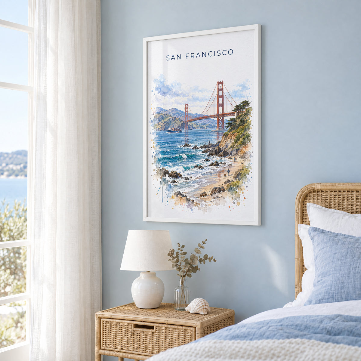 Golden Gate Bridge watercolor print in a white frame on a pale blue wall beside a rattan bed in a coastal bedroom