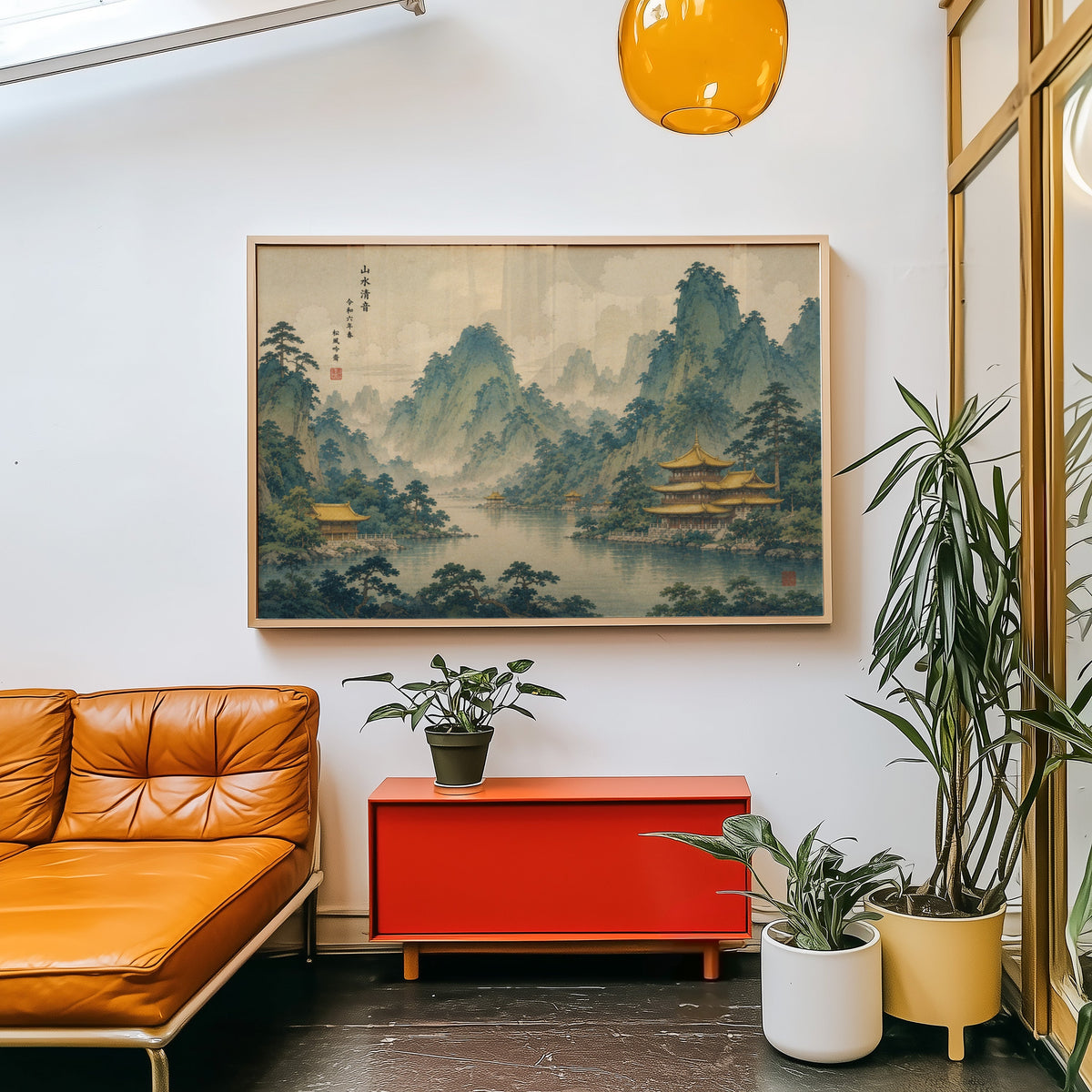 Golden temples below green mountains print in a light wood frame above a red cabinet beside a tan leather daybed