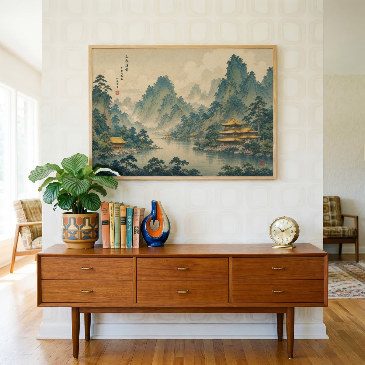 Golden temples below green mountains print in a light oak frame above a teak credenza with books and a brass clock