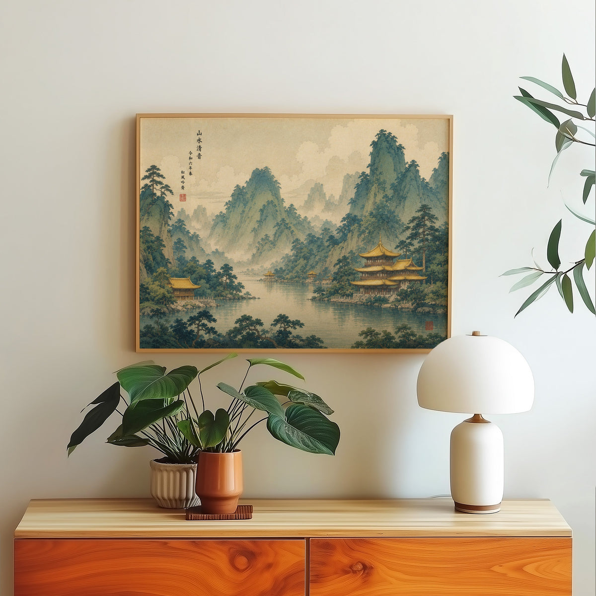 Golden temples below green mountains print in a light oak frame above a wood sideboard with a white dome lamp