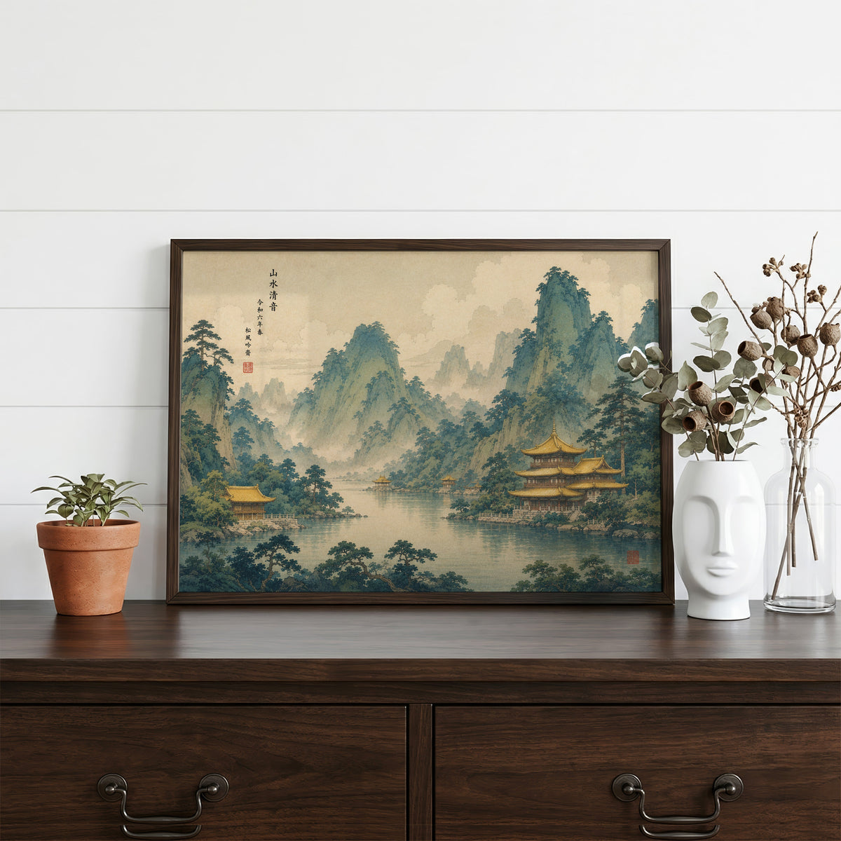 Golden temples below green mountains print in a dark walnut frame on a dark wood dresser beside a white face vase