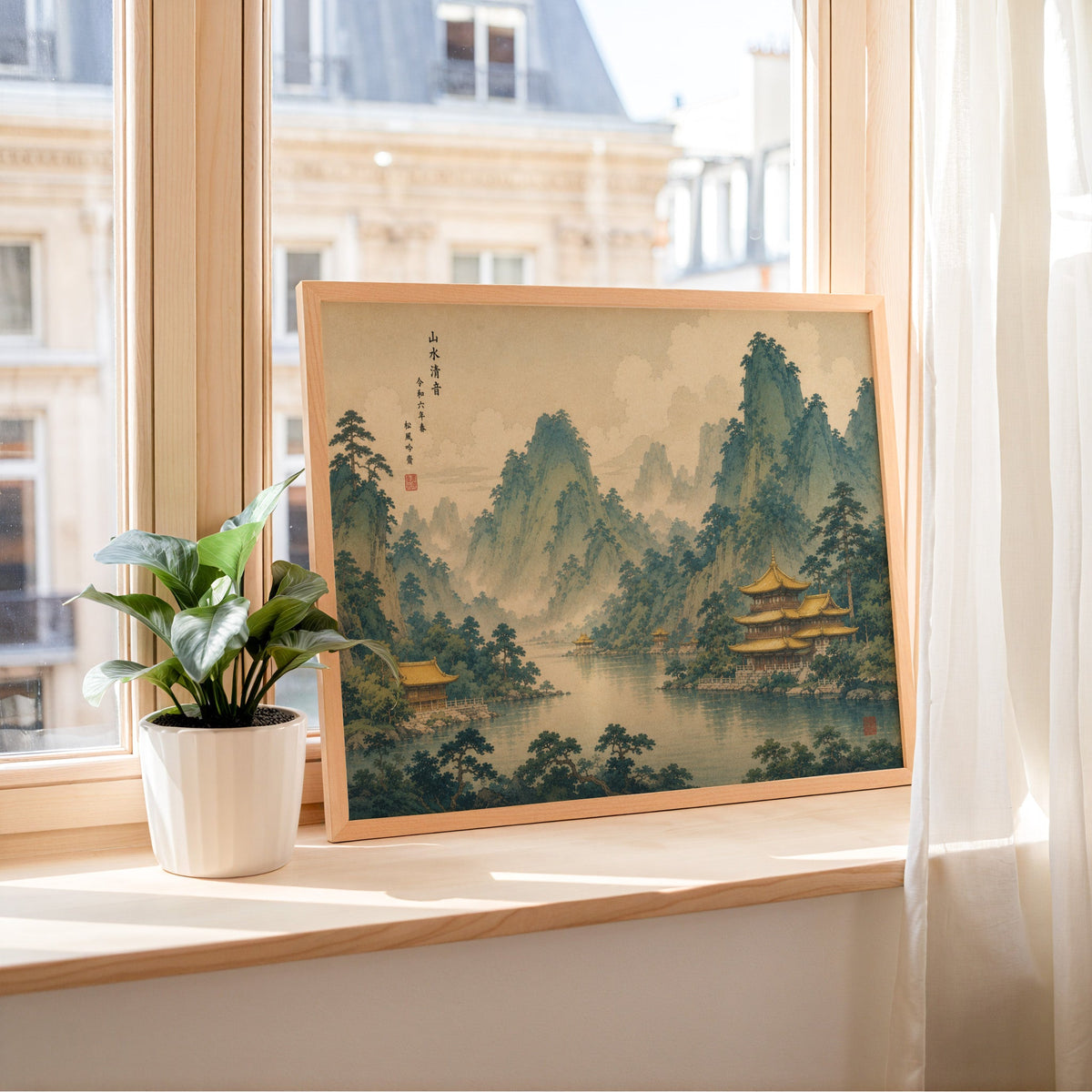 Golden temples below green mountains print in a light wood frame leaning on a sunny windowsill by sheer curtains