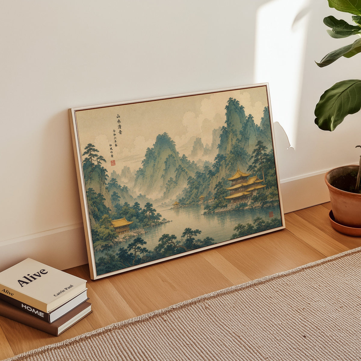 Golden temples below green mountains print in a white floater frame leaning on a wood floor beside stacked books