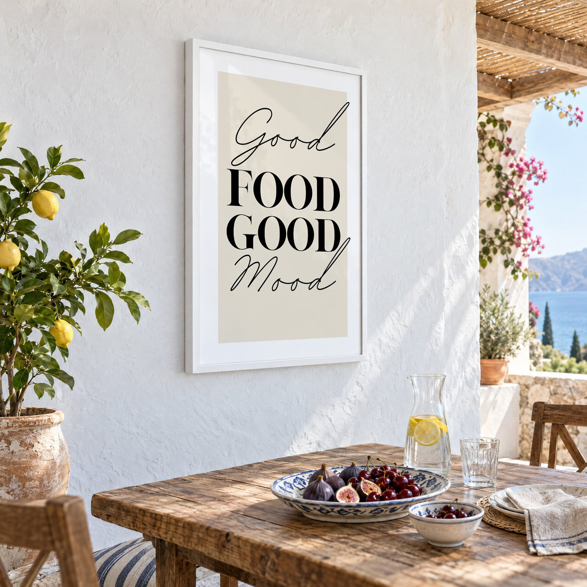 Good FOOD GOOD Mood serif script print in a white frame on a whitewashed terrace wall above a lunch table with figs and cherries, seen at an angle