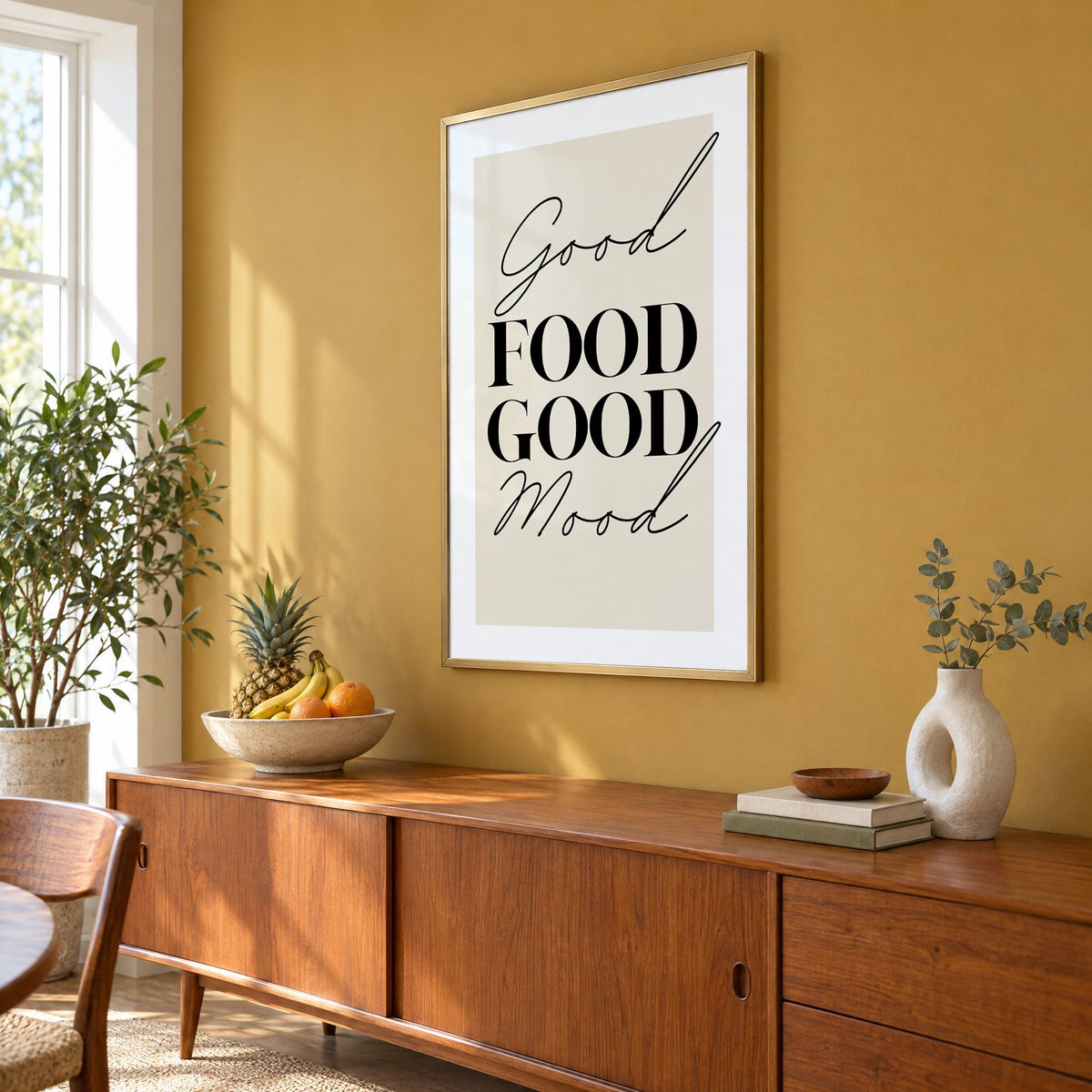 Good FOOD GOOD Mood serif script print in a brass frame on a mustard wall above a teak sideboard with a fruit bowl, seen at an angle