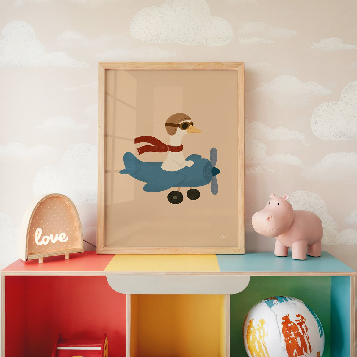 Goose pilot in blue airplane nursery print in an oak frame on a rainbow toy shelf against cloud wallpaper