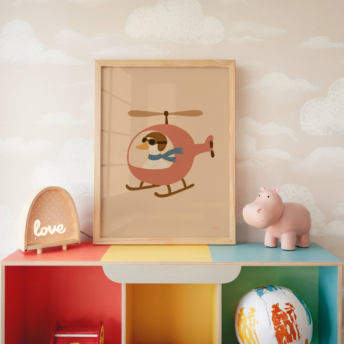 Goose pilot in pink helicopter nursery print in an oak frame on a rainbow toy shelf against cloud wallpaper