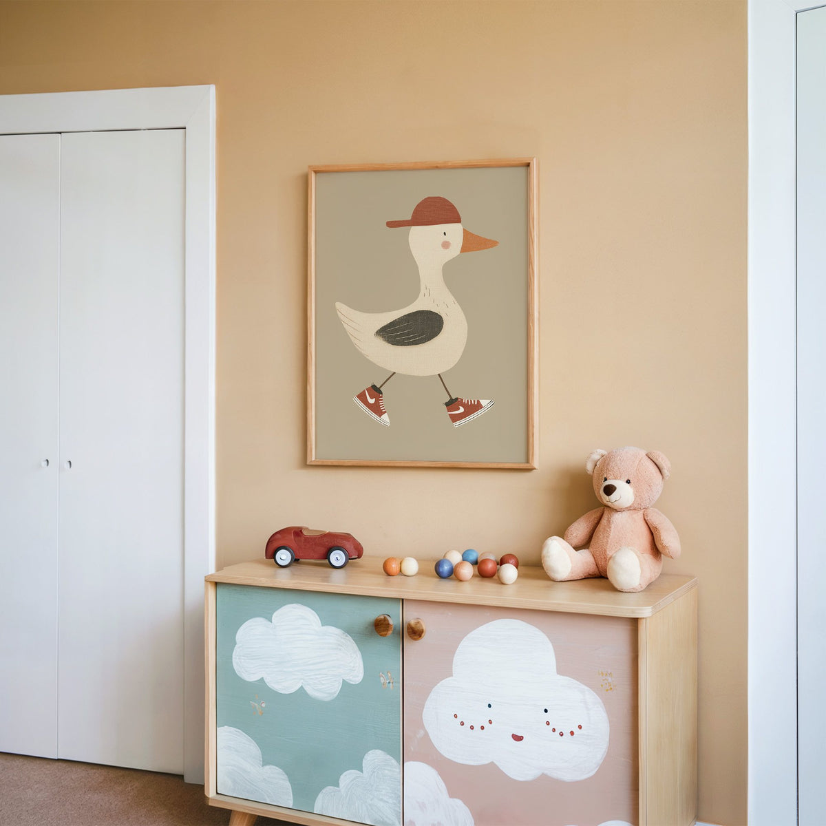 Goose red cap and sneakers nursery print in an oak frame on a peach wall above a cloud-painted cabinet