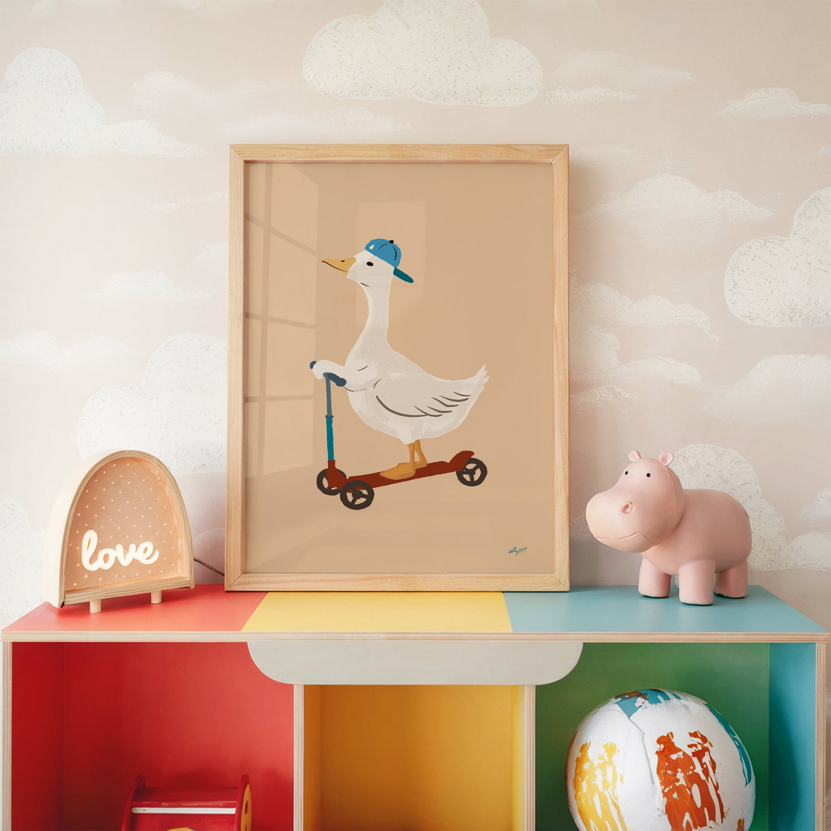 Goose riding scooter in blue cap nursery print in an oak frame on a rainbow toy shelf against cloud wallpaper
