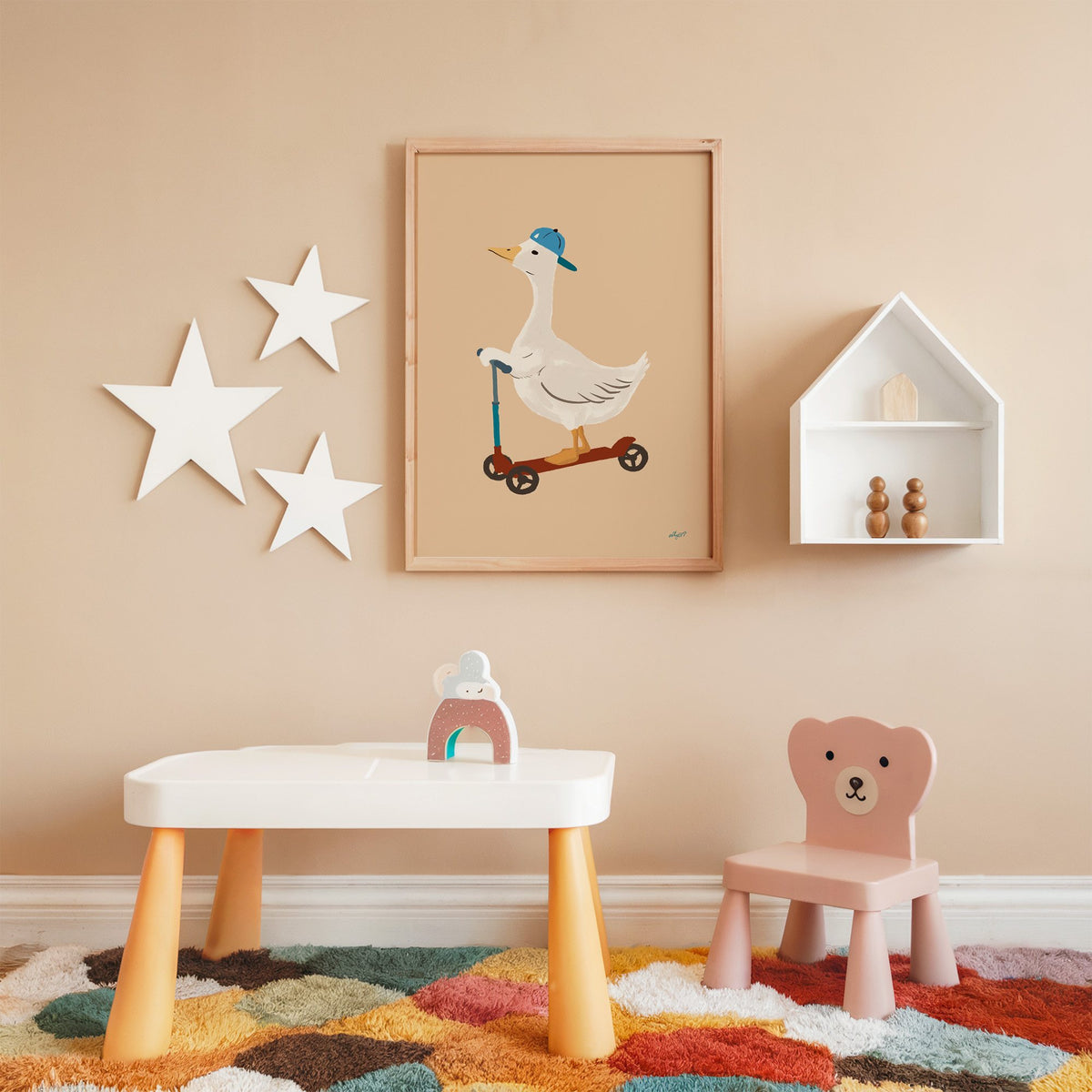 Goose riding scooter in blue cap nursery print in an oak frame on a peach wall above a kids table with wall stars