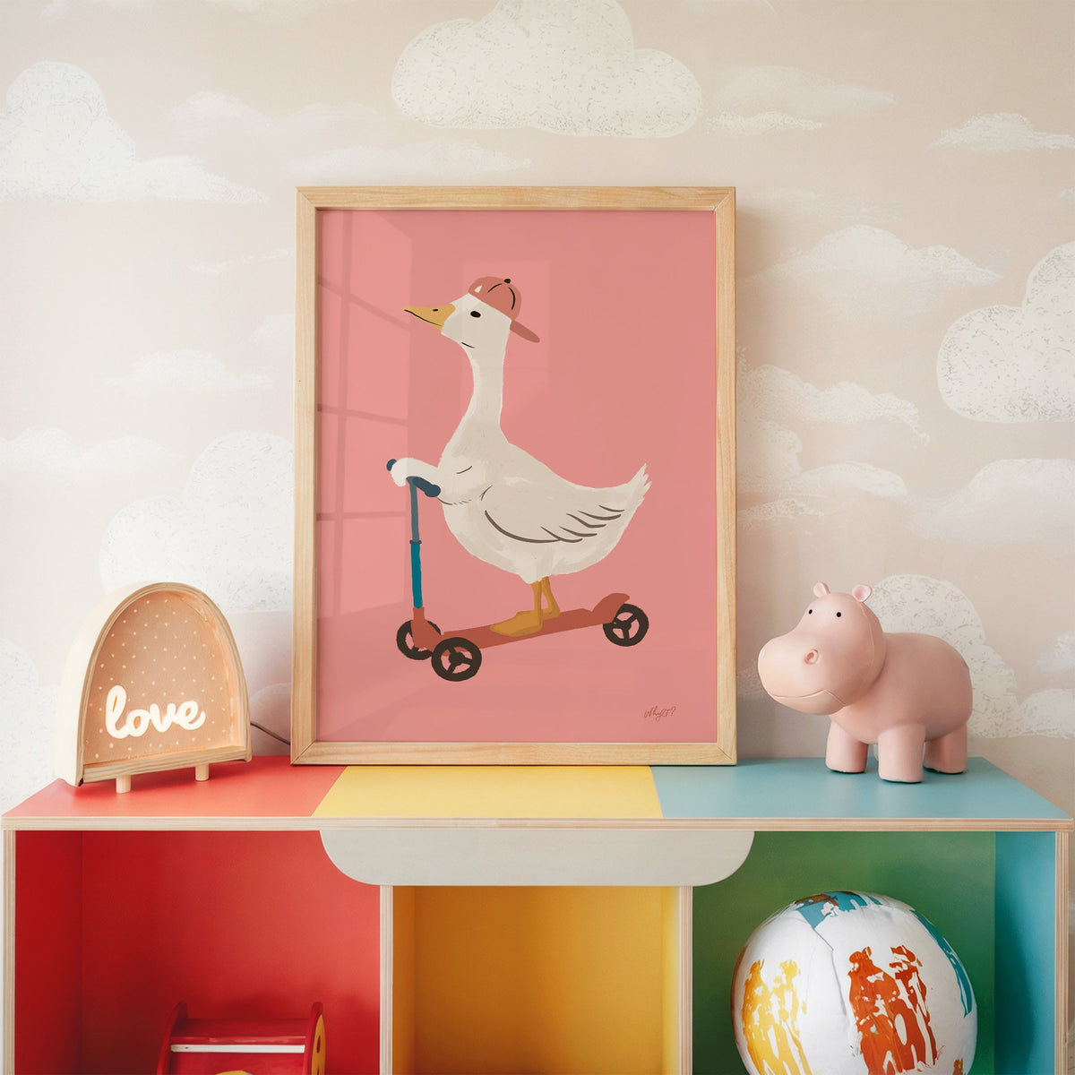 Goose on scooter in pink cap nursery print in an oak frame on a rainbow toy shelf against cloud wallpaper