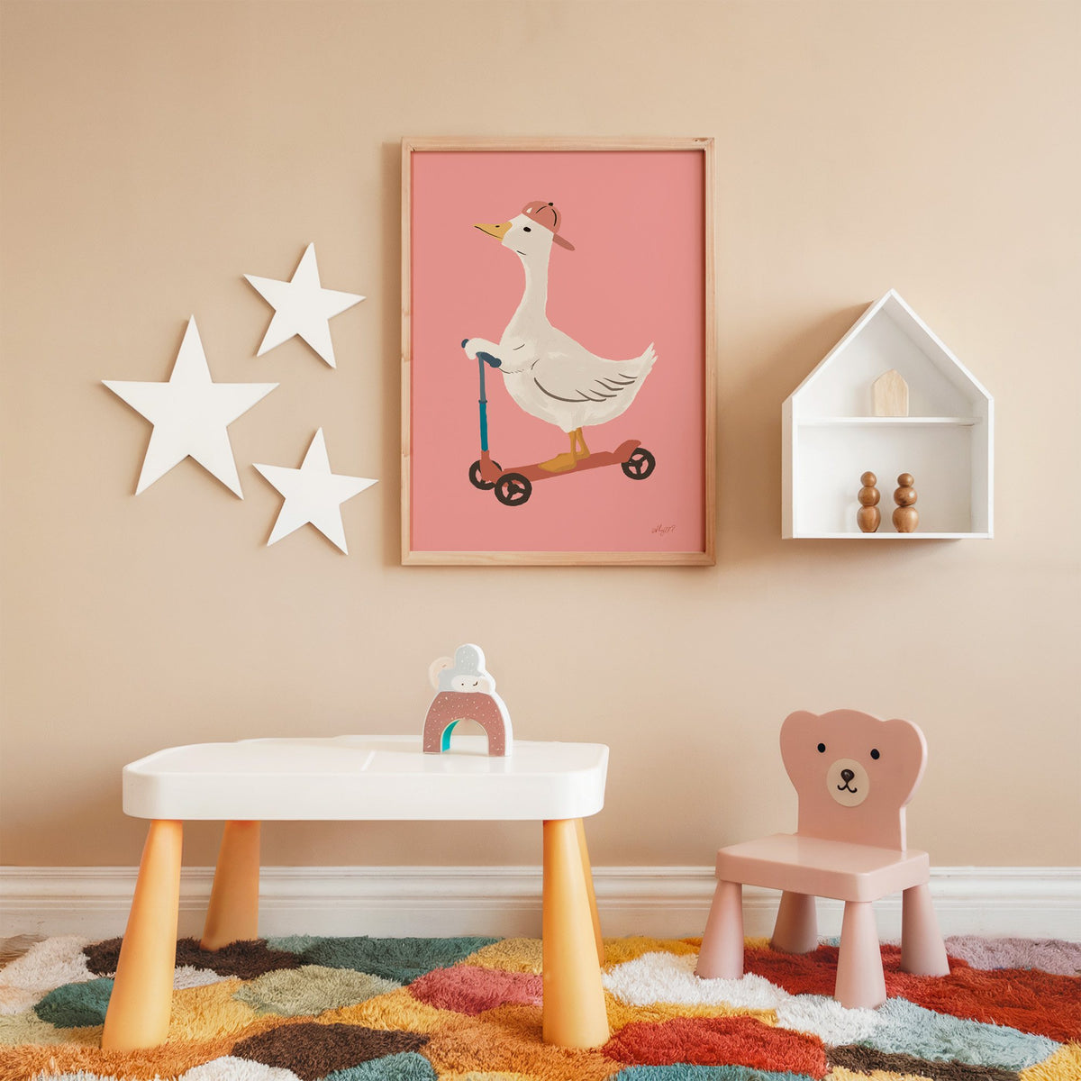 Goose on scooter in pink cap nursery print in an oak frame on a peach wall above a kids table with wall stars