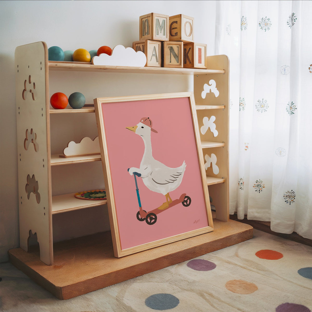 Goose on scooter in pink cap nursery print in an oak frame leaning on a toy shelf with wooden letter blocks