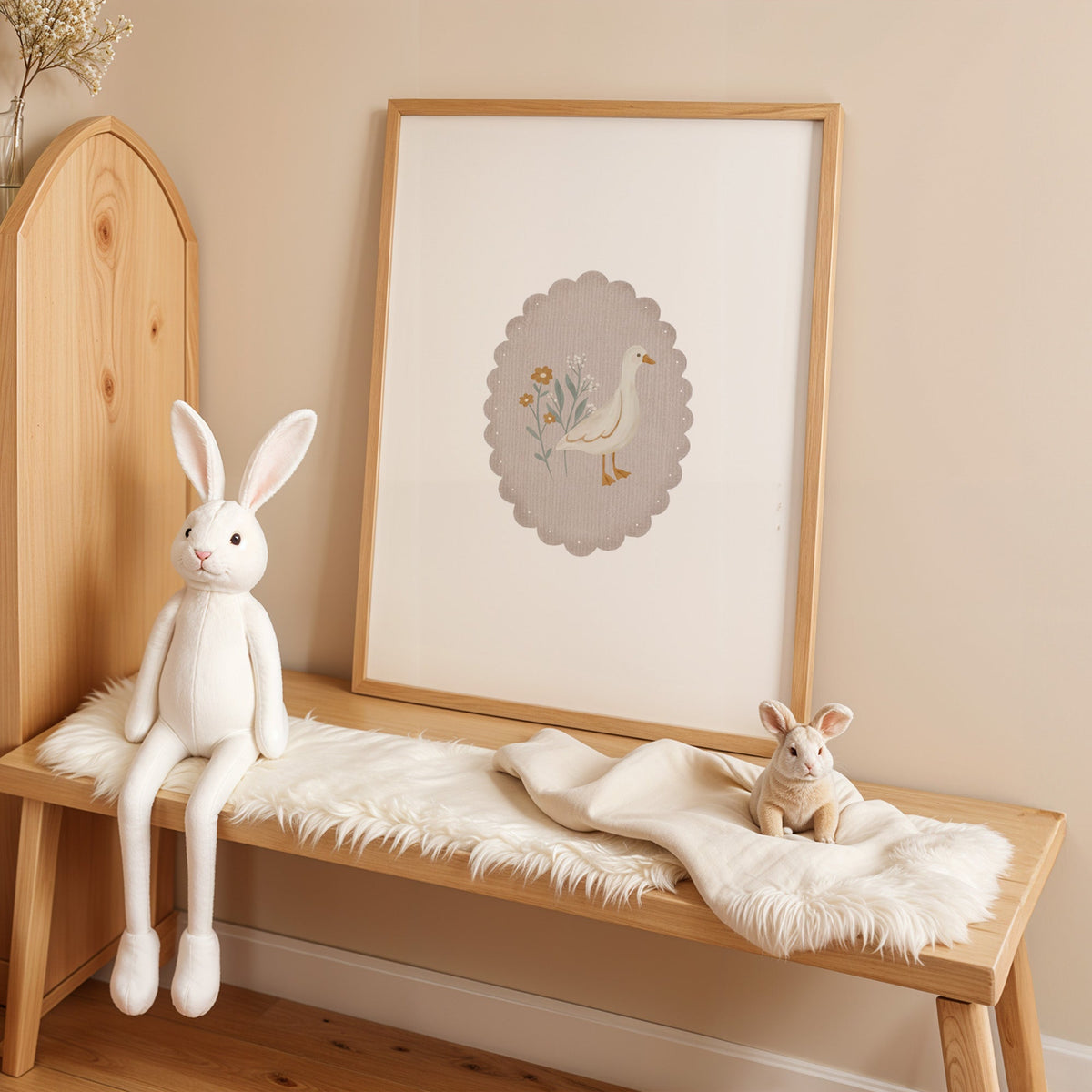 White goose and wildflowers in scalloped oval nursery print in an oak frame on a sheepskin bench beside a white plush bunny