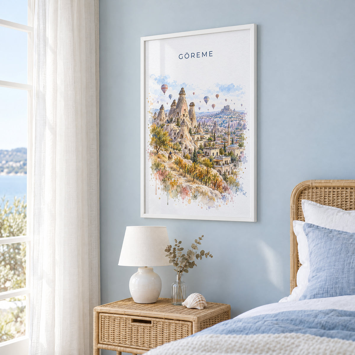 Göreme watercolor print in a white frame on a pale blue wall beside a rattan bed in a coastal bedroom
