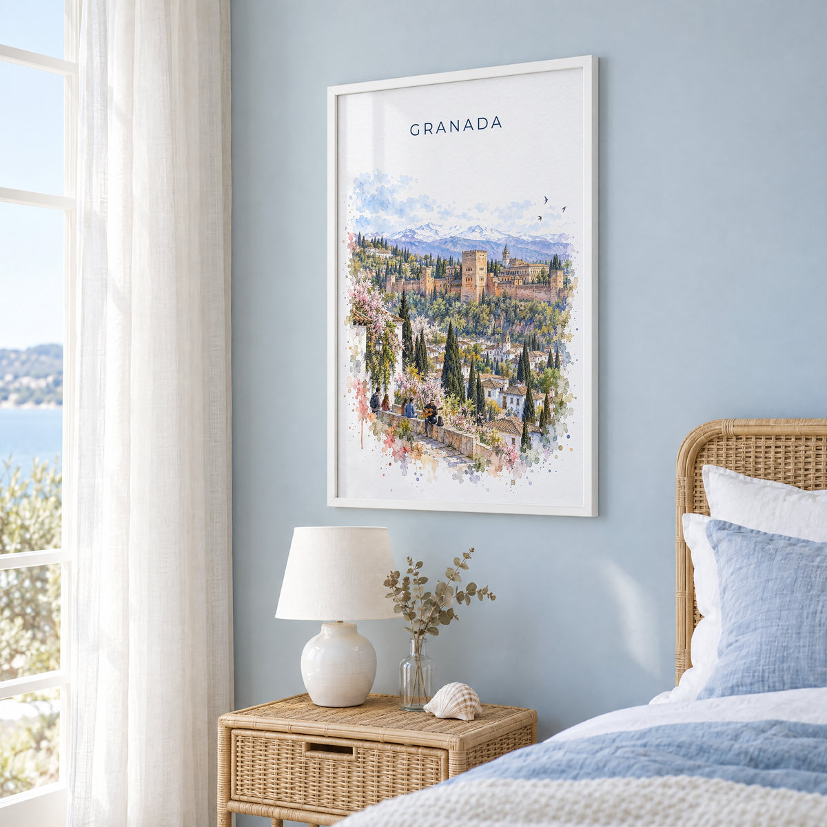 Granada, Spain watercolor print in a white frame on a pale blue wall beside a rattan bed in a coastal bedroom