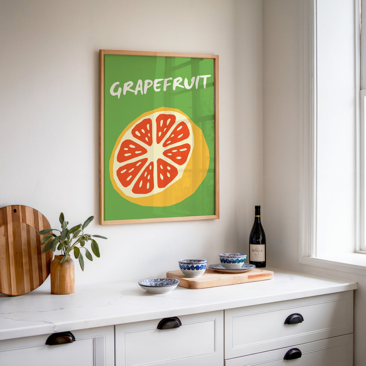 Grapefruit half print in an oak frame above a white sideboard with a bottle of wine