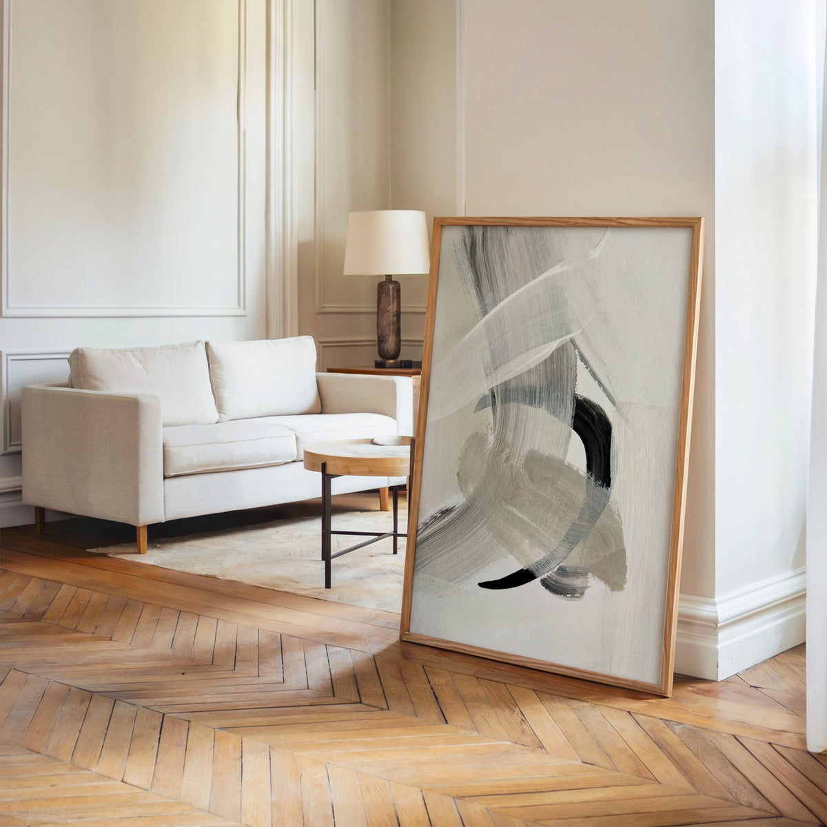 Gray and beige brushstrokes with black arc print in an oak frame leaning on a herringbone floor by a cream linen sofa