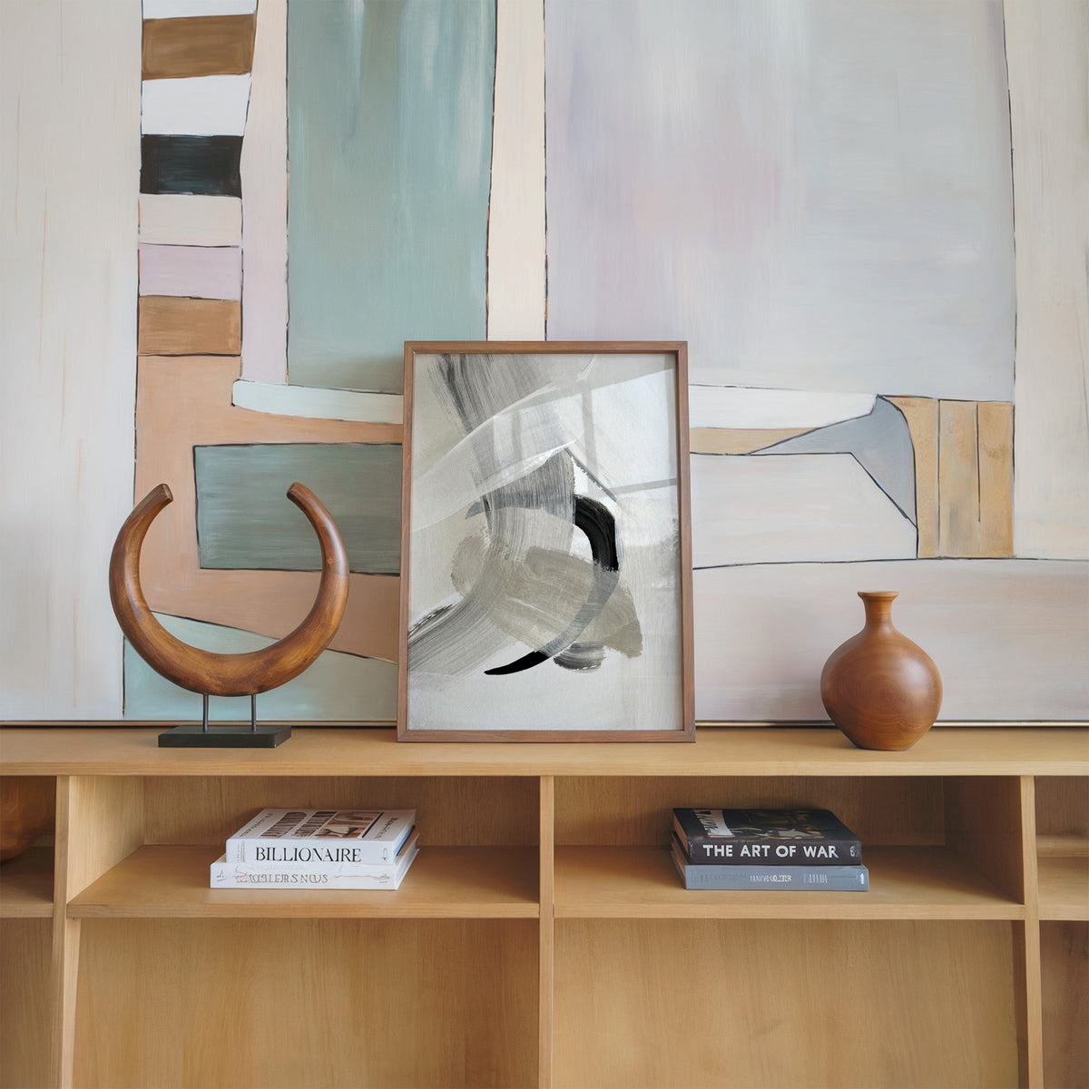 Gray and beige brushstrokes with black arc print in a walnut frame on an oak shelf beside a wooden crescent sculpture