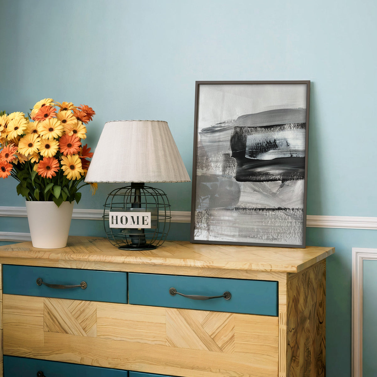 Gray and black dry brush layers abstract print in a charcoal frame on a teal dresser beside orange and yellow daisies