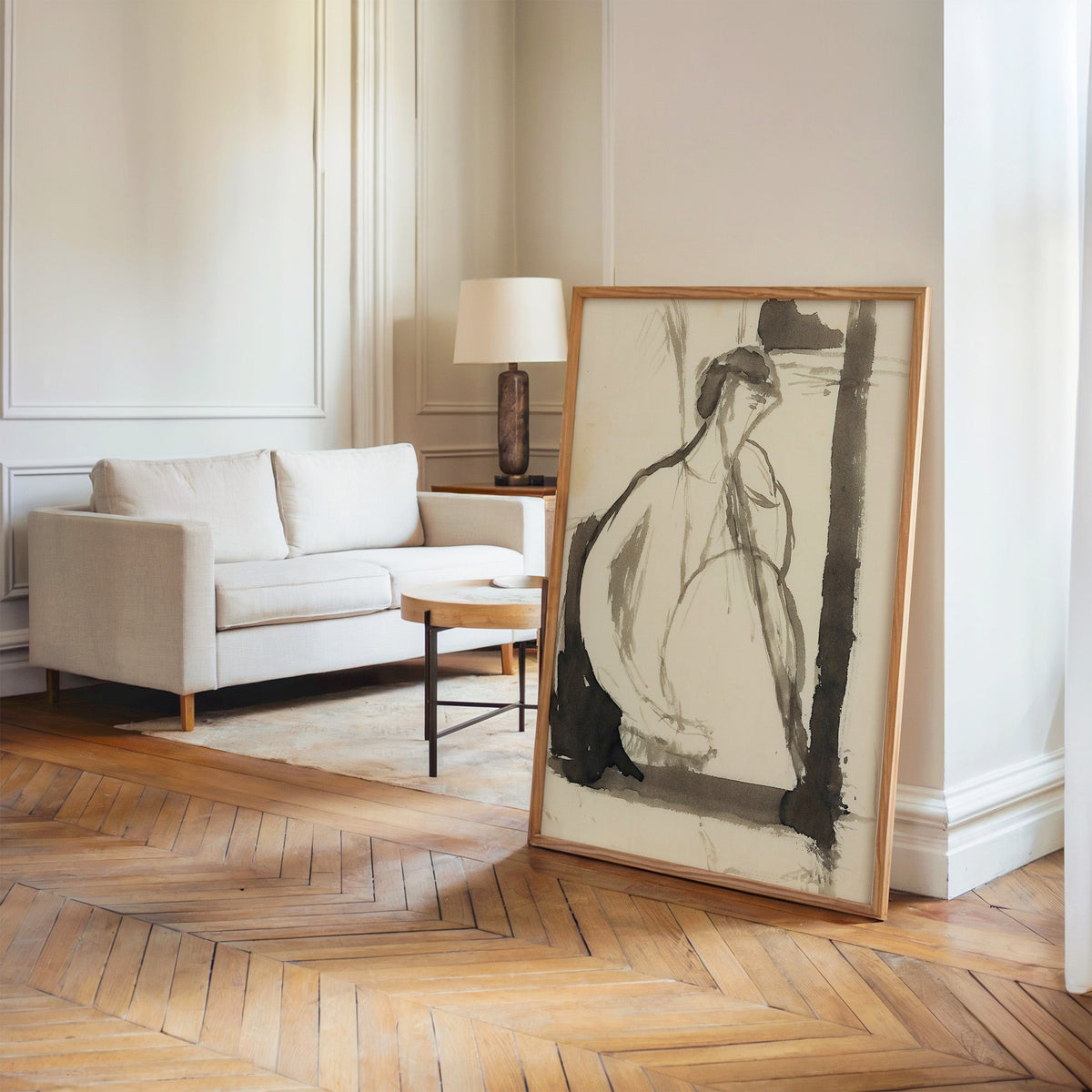 Gray ink seated woman figure sketch print in an oak frame leaning on a herringbone floor by a cream linen sofa