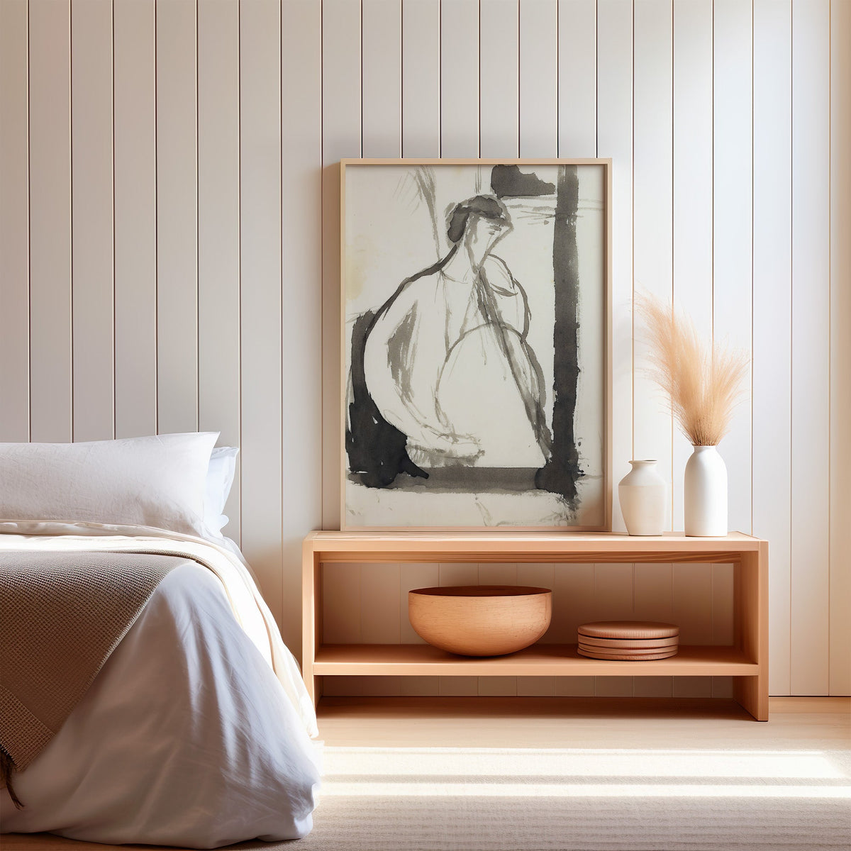 Gray ink seated woman figure sketch print in a light oak frame on a wood bench by a bed with pampas grass