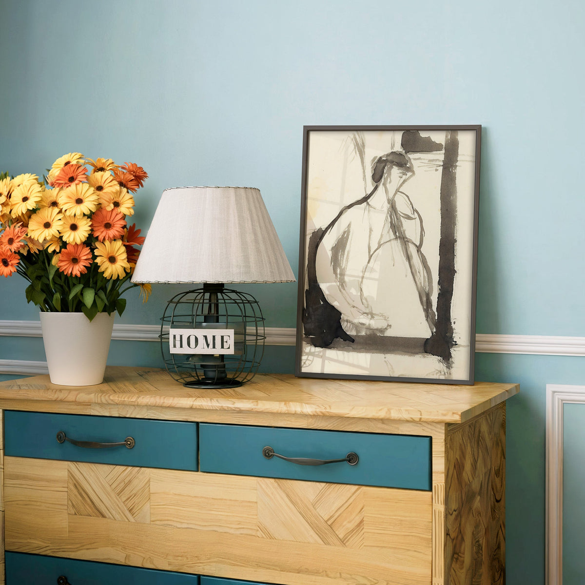 Gray ink seated woman figure sketch print in a charcoal frame on a teal dresser beside orange and yellow daisies