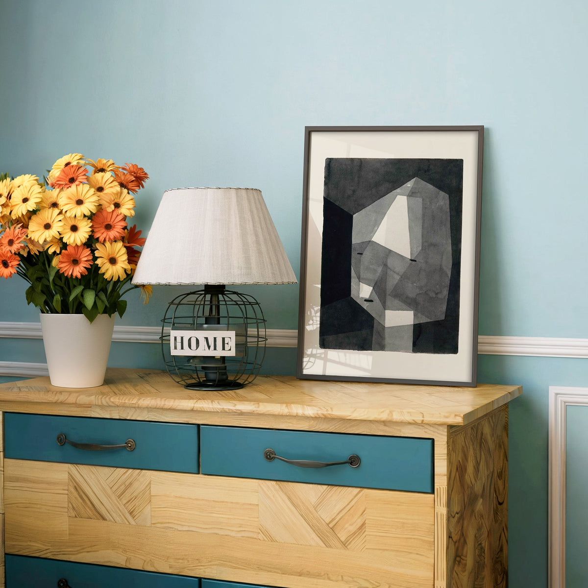 Gray faceted crystal head watercolor abstract print in a charcoal frame on a teal dresser beside orange and yellow daisies