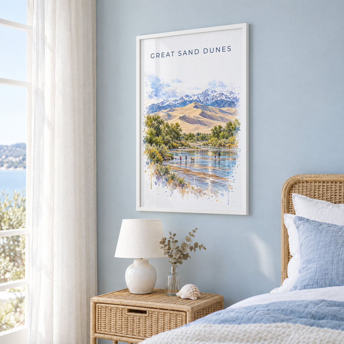Great Sand Dunes National Park watercolor print in a white frame on a pale blue wall beside a rattan bed in a coastal bedroom