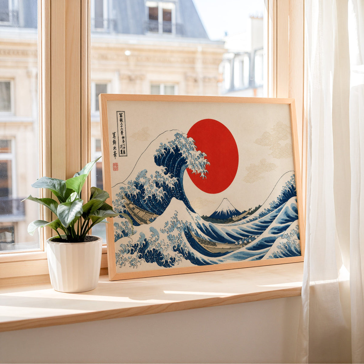 Great Wave with a red sun print in a light wood frame leaning on a sunny windowsill by sheer curtains