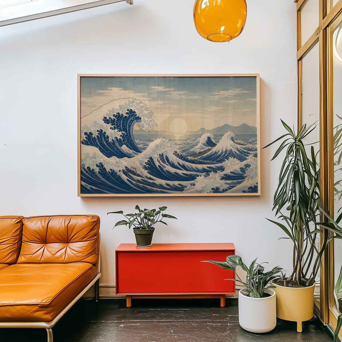 Great wave at sunrise over the sea print in a light wood frame above a red cabinet beside a tan leather daybed
