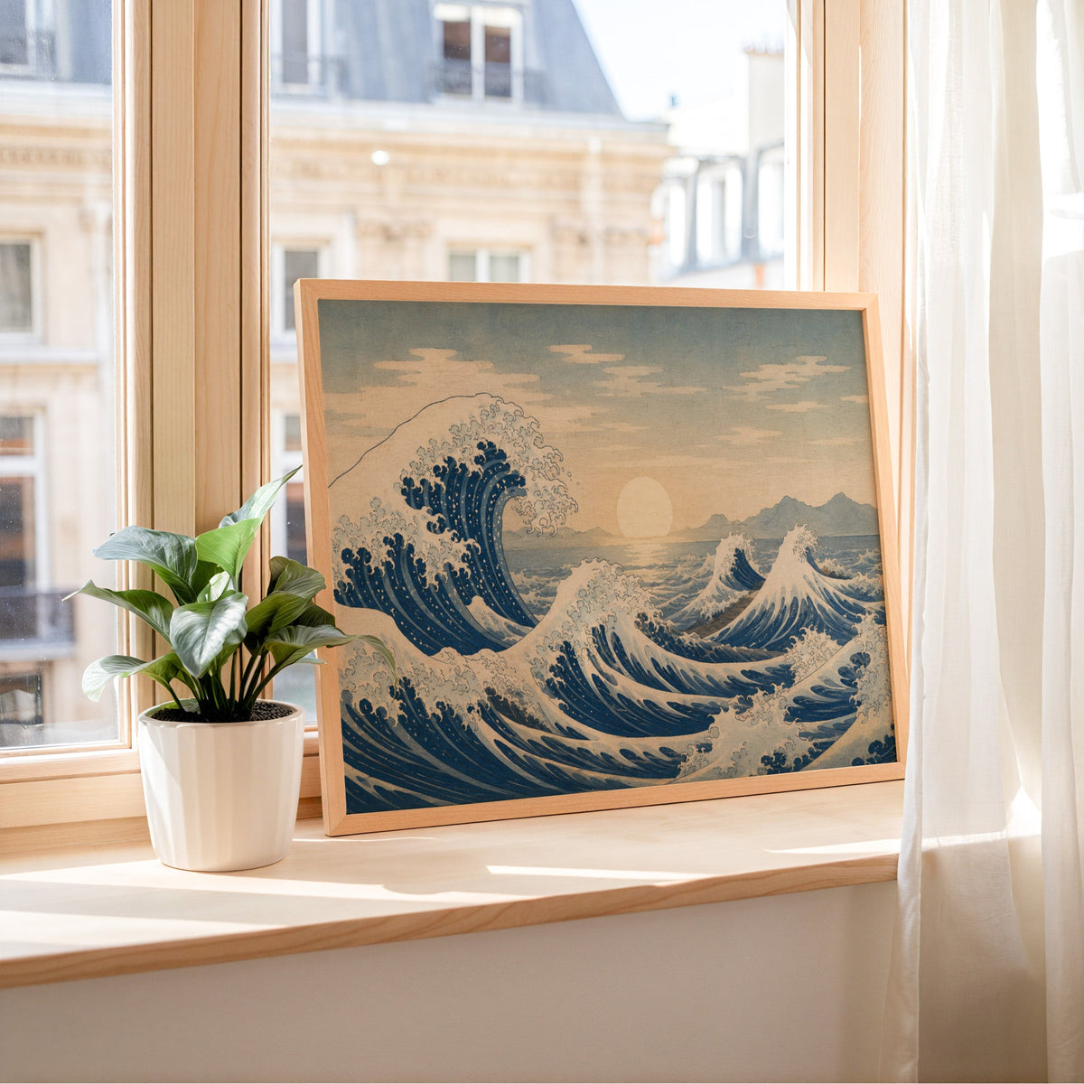 Great wave at sunrise over the sea print in a light wood frame leaning on a sunny windowsill by sheer curtains