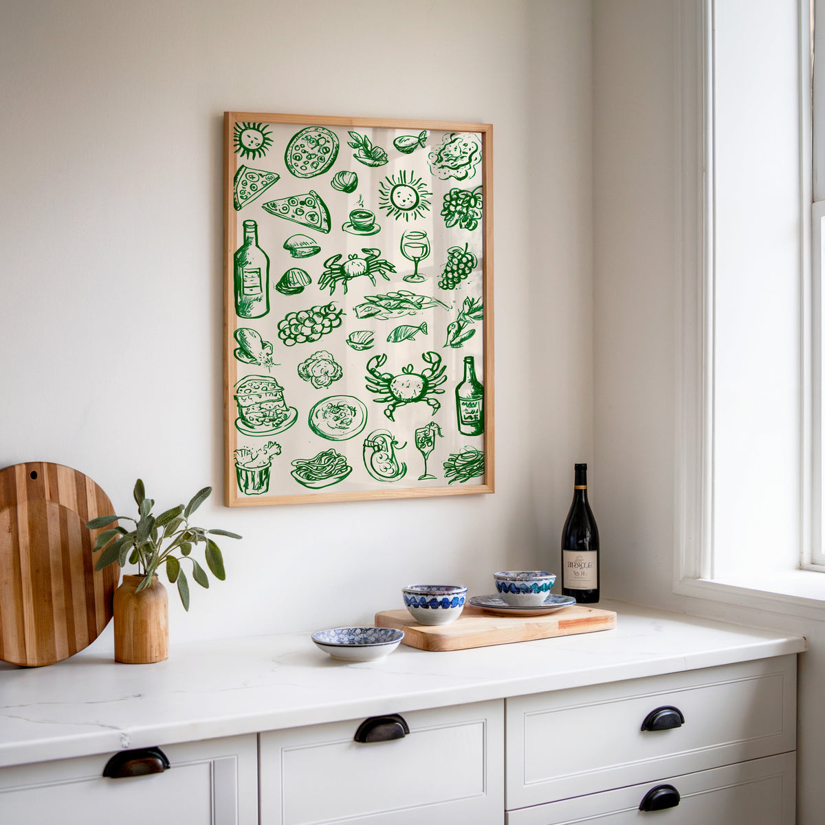 Green food doodle print in an oak frame above a white sideboard with a bottle of wine