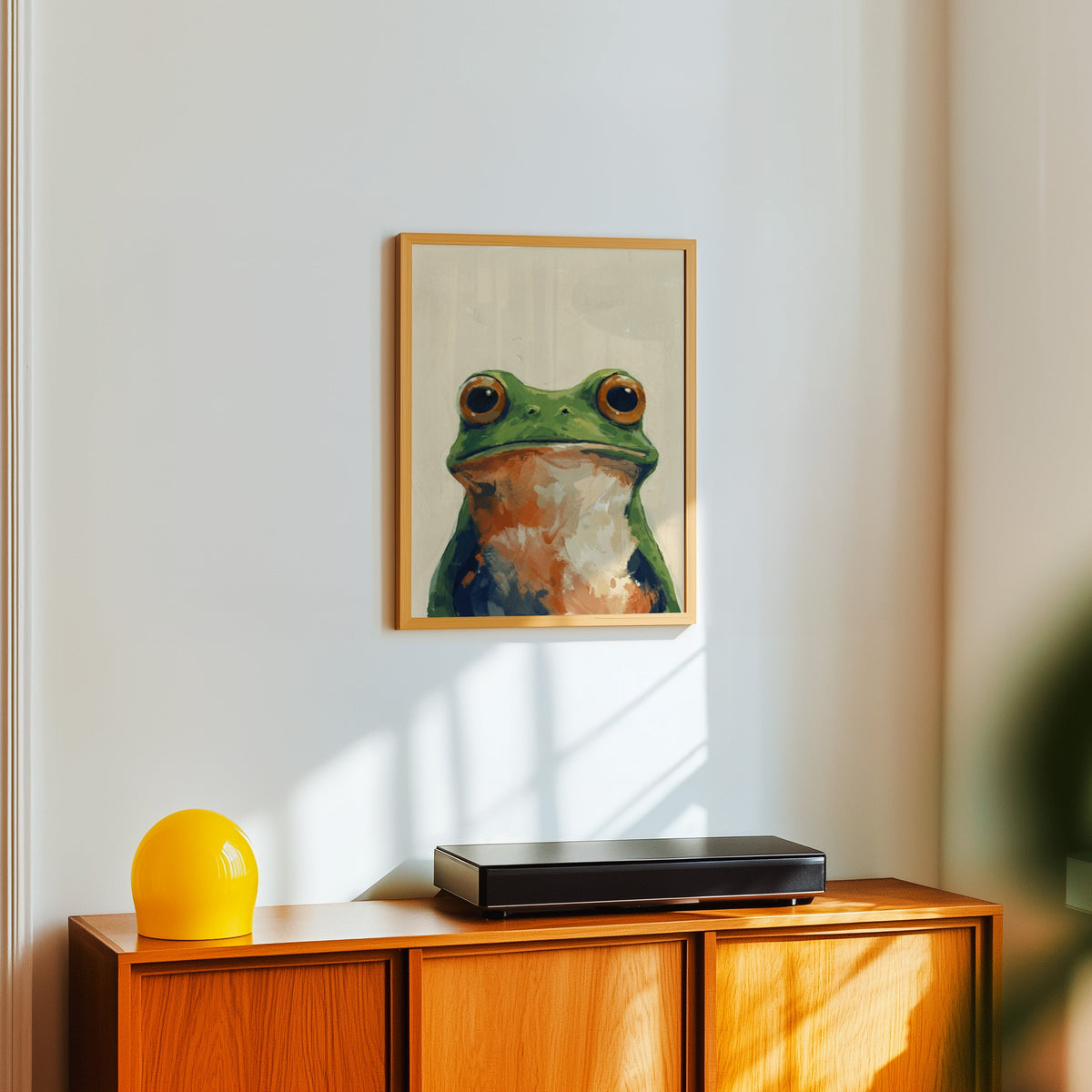 Cream and green frog print in a light oak frame above a teak sideboard with a yellow lamp