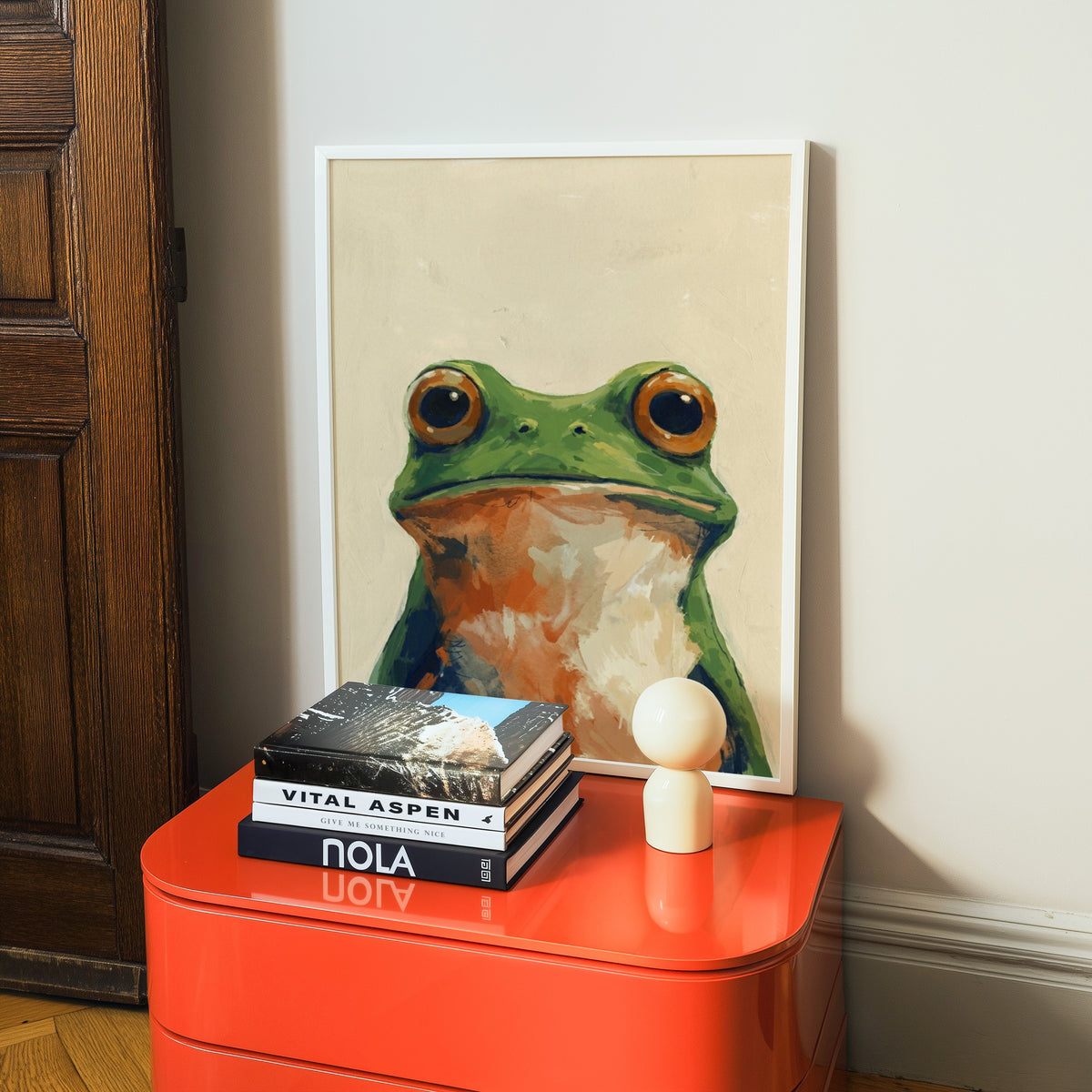 Funny frog print in a white frame on a glossy orange cabinet with books
