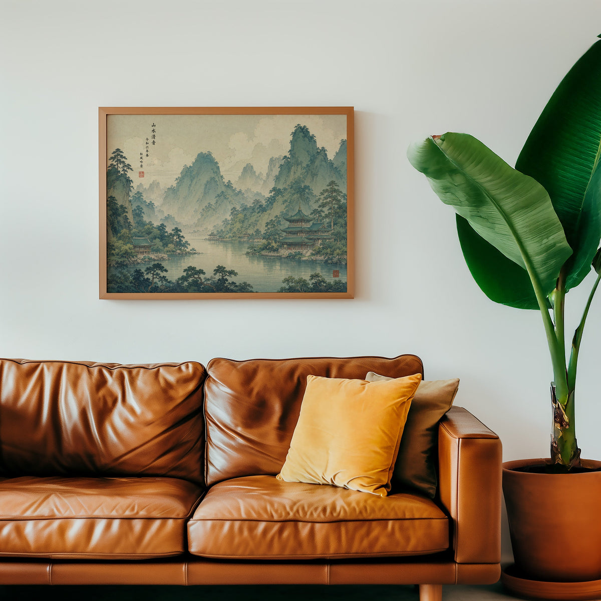 Green mountain lake with temples print in a warm oak frame above a cognac leather sofa with a mustard pillow