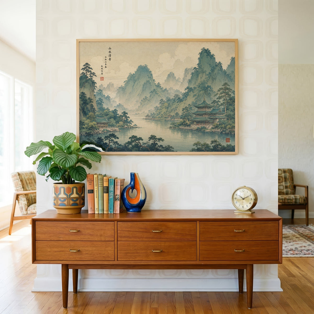 Green mountain lake with temples print in a light oak frame above a teak credenza with books and a brass clock