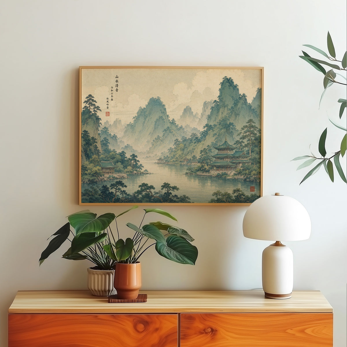 Green mountain lake with temples print in a light oak frame above a wood sideboard with a white dome lamp