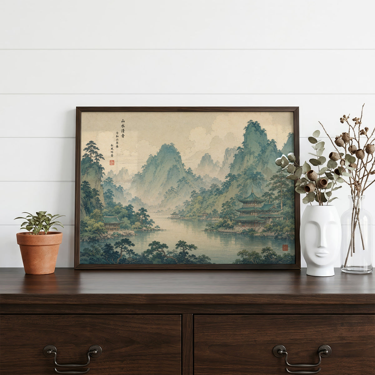 Green mountain lake with temples print in a dark walnut frame on a dark wood dresser beside a white face vase