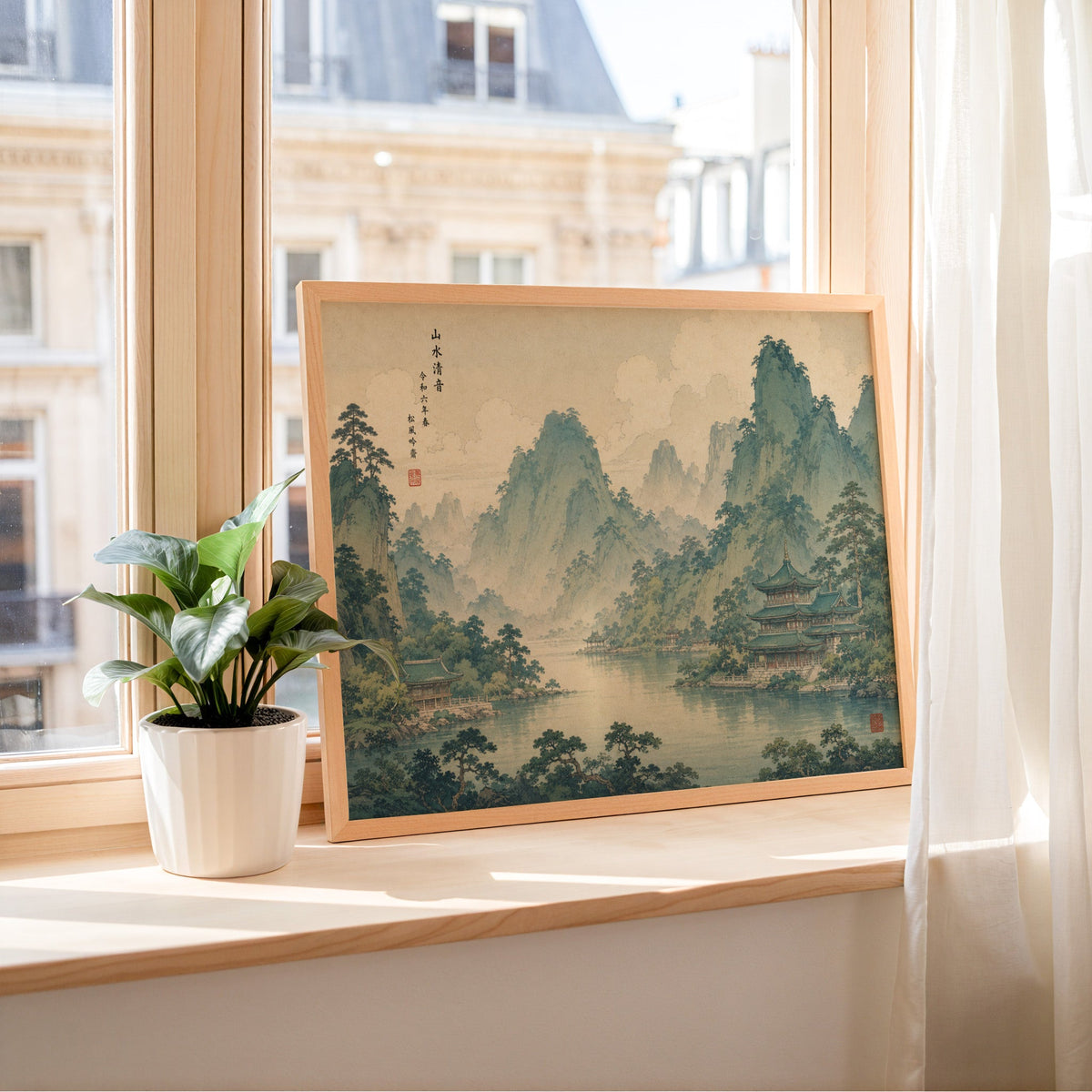 Green mountain lake with temples print in a light wood frame leaning on a sunny windowsill by sheer curtains
