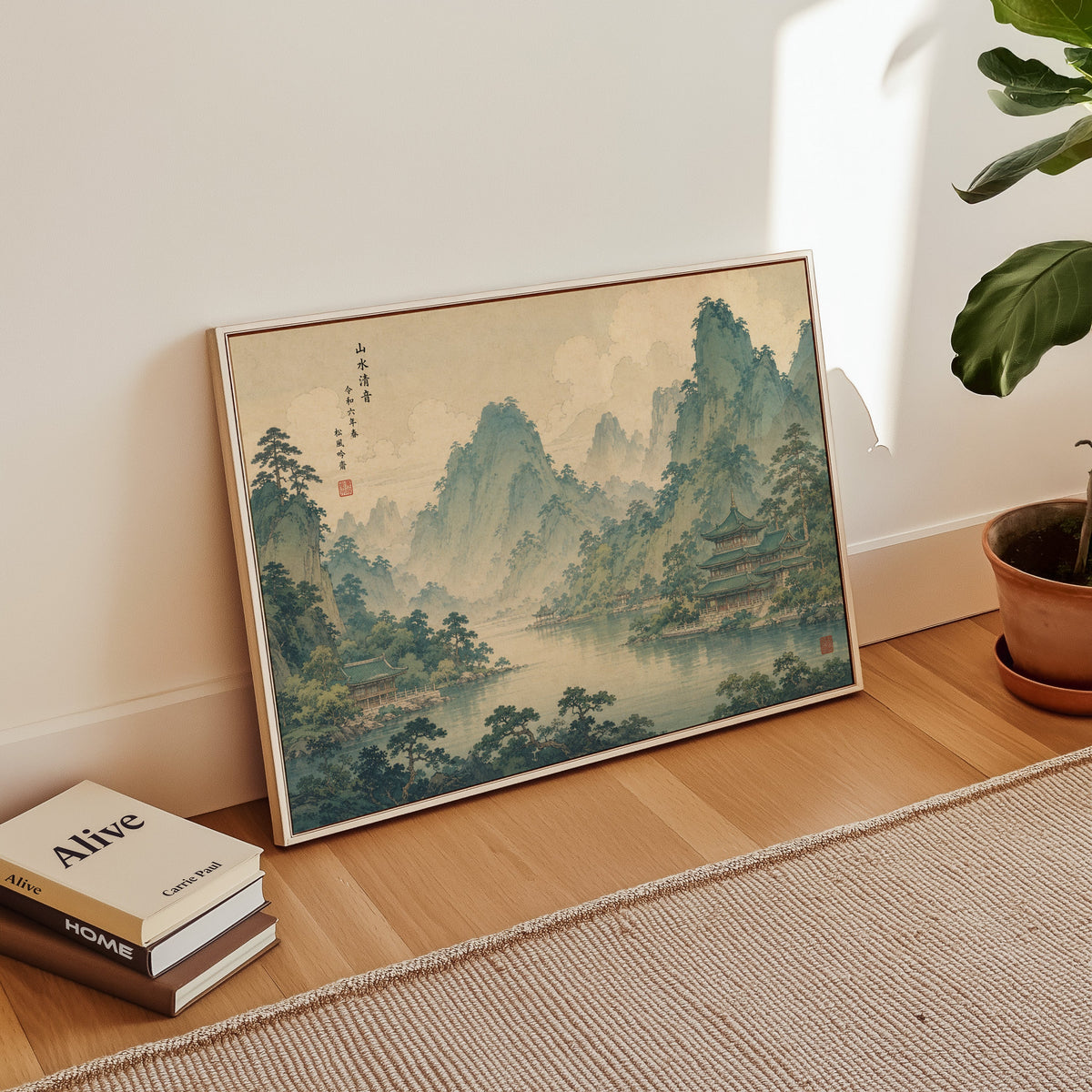 Green mountain lake with temples print in a white floater frame leaning on a wood floor beside stacked books