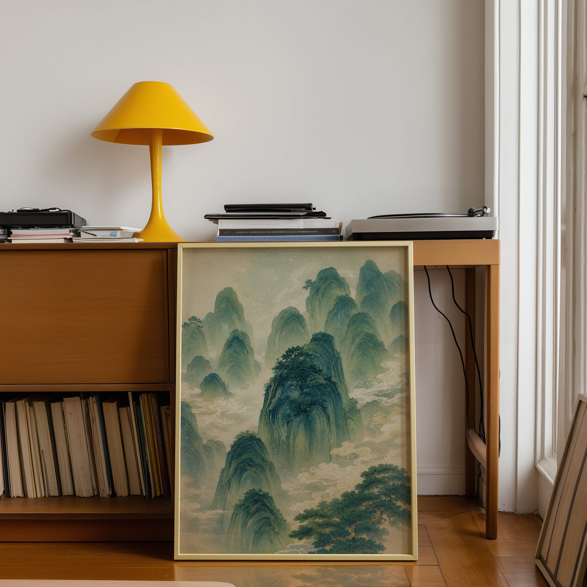 green mountains and clouds landscape print shown in a gold frame leaning under a teak desk with a yellow lamp