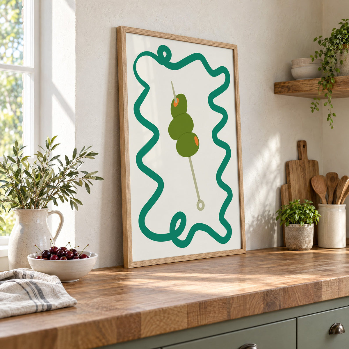 Green olives cocktail pick print in an oak frame leaning on a butcher block island beside a bowl of cherries