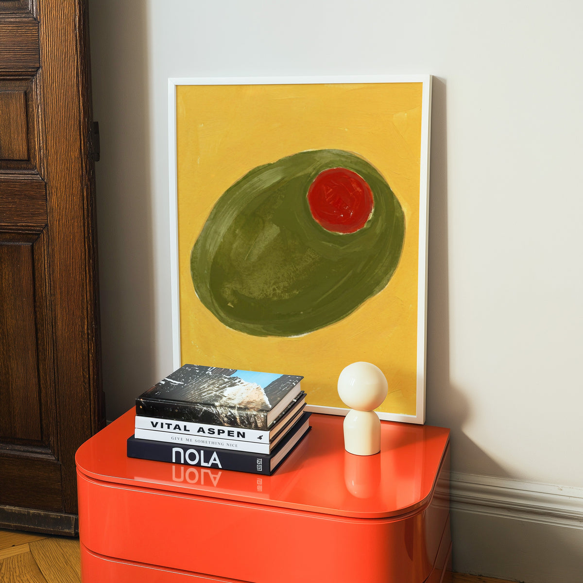 Pimiento olive kitchen print in a white frame on a glossy orange cabinet with books