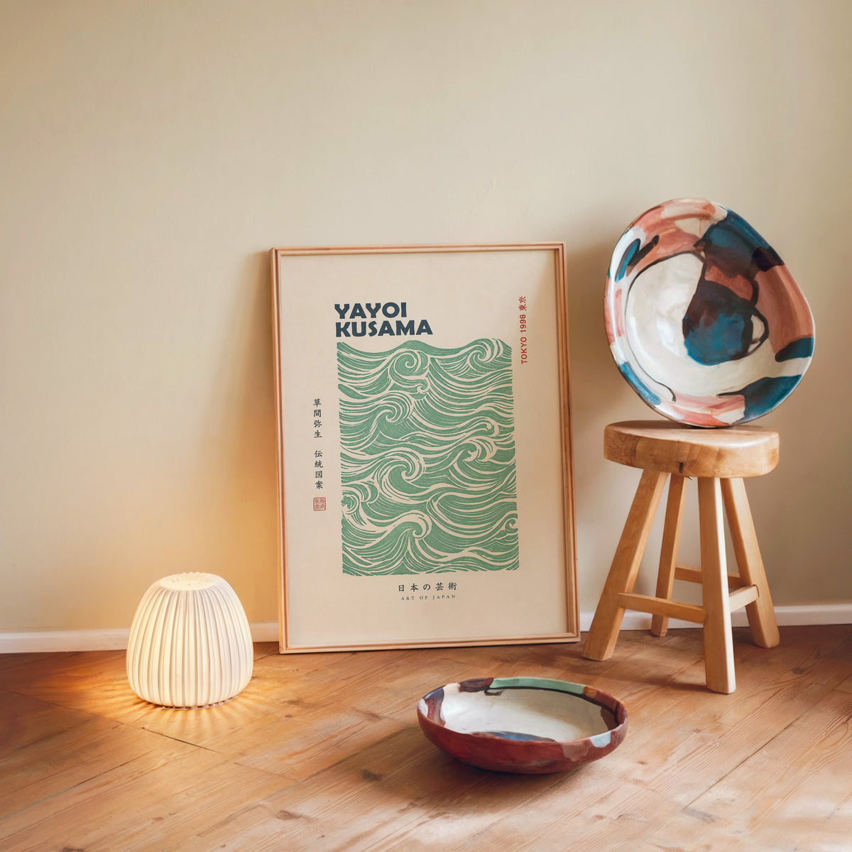 green wave pattern with bold type print in a light wood frame leaning on a wooden floor by a glowing lamp and stool