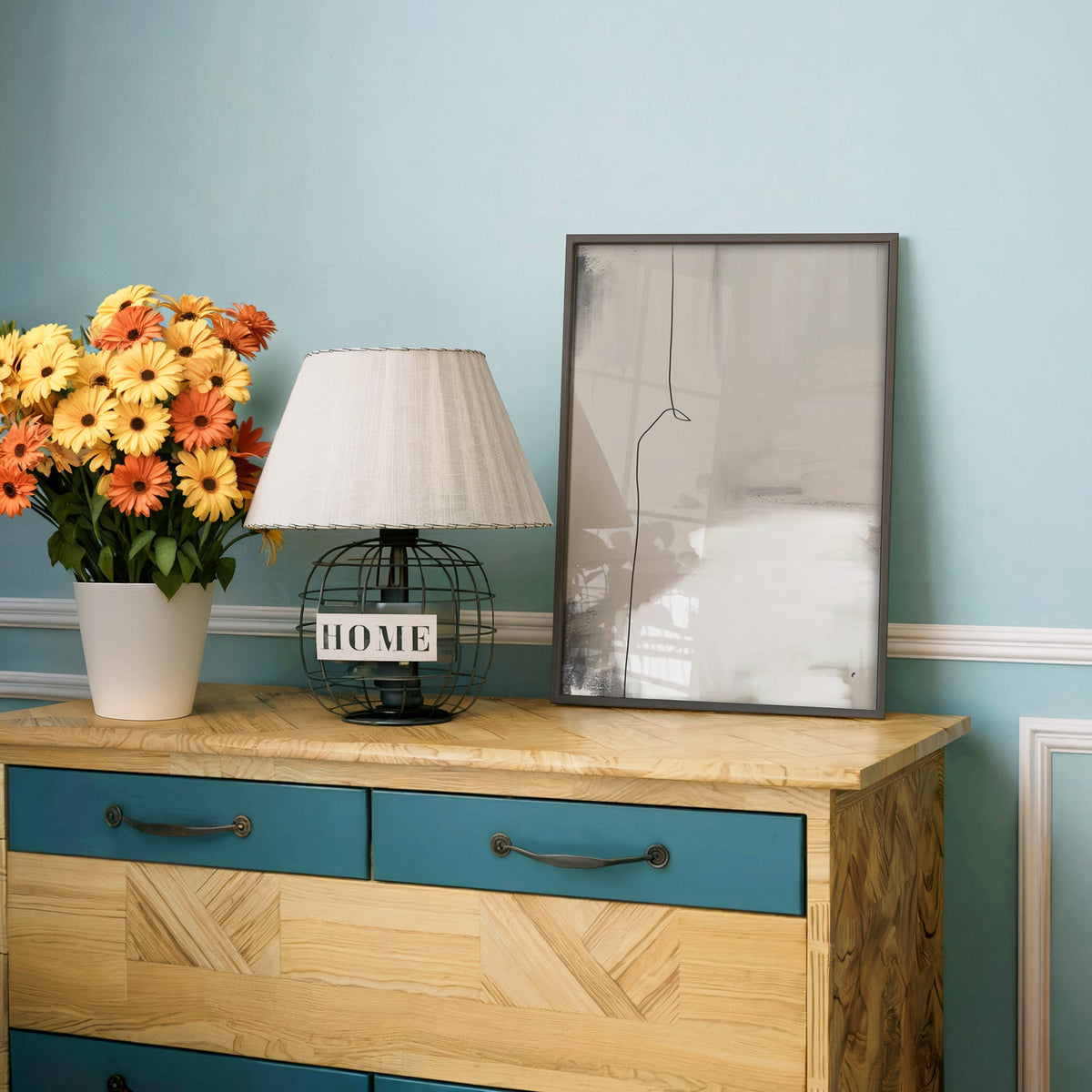 Greige wash with single thin line print in a charcoal frame on a teal dresser beside orange and yellow daisies