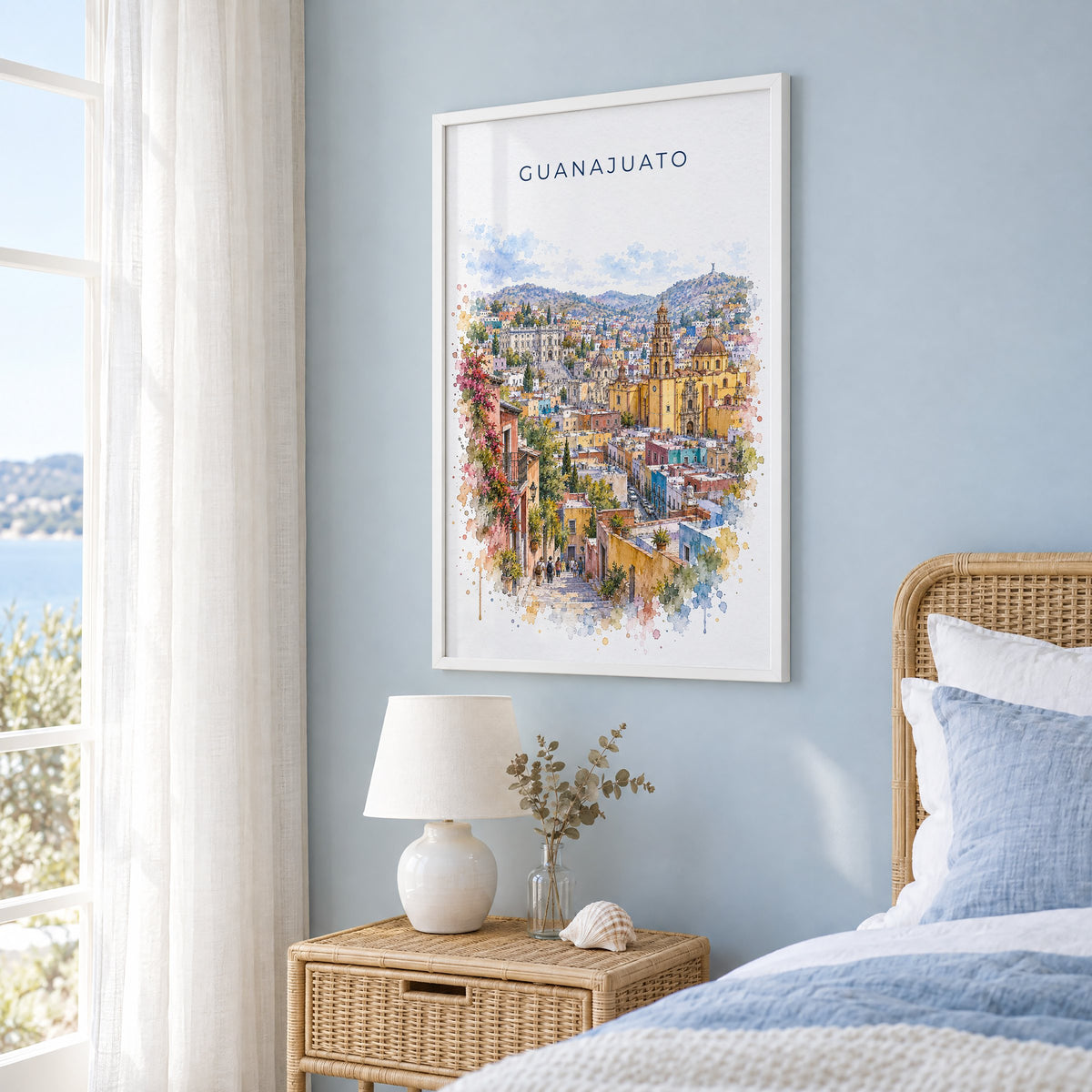 Guanajuato watercolor print in a white frame on a pale blue wall beside a rattan bed in a coastal bedroom
