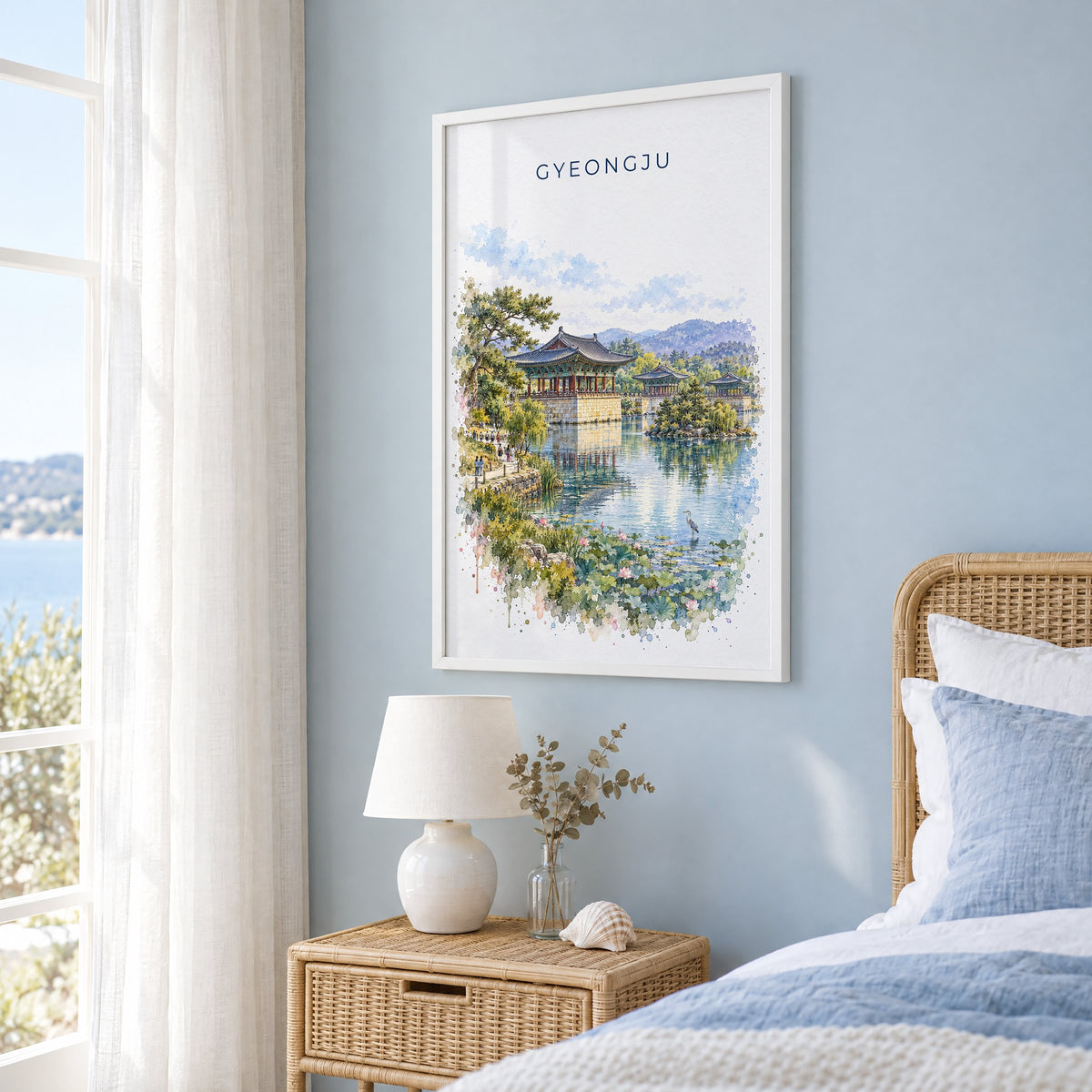 Gyeongju watercolor print in a white frame on a pale blue wall beside a rattan bed in a coastal bedroom