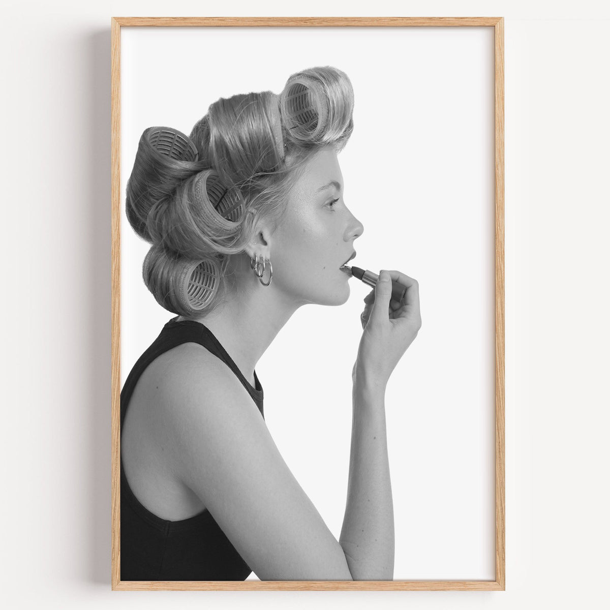 Woman in hair rollers applying lipstick profile print in a light oak frame on a plain white wall