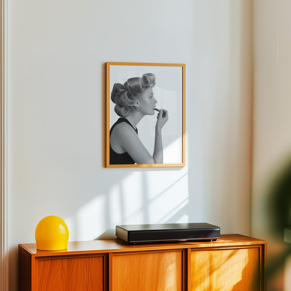 Woman in hair rollers applying lipstick profile print in an oak frame above a teak sideboard with a yellow glass lamp